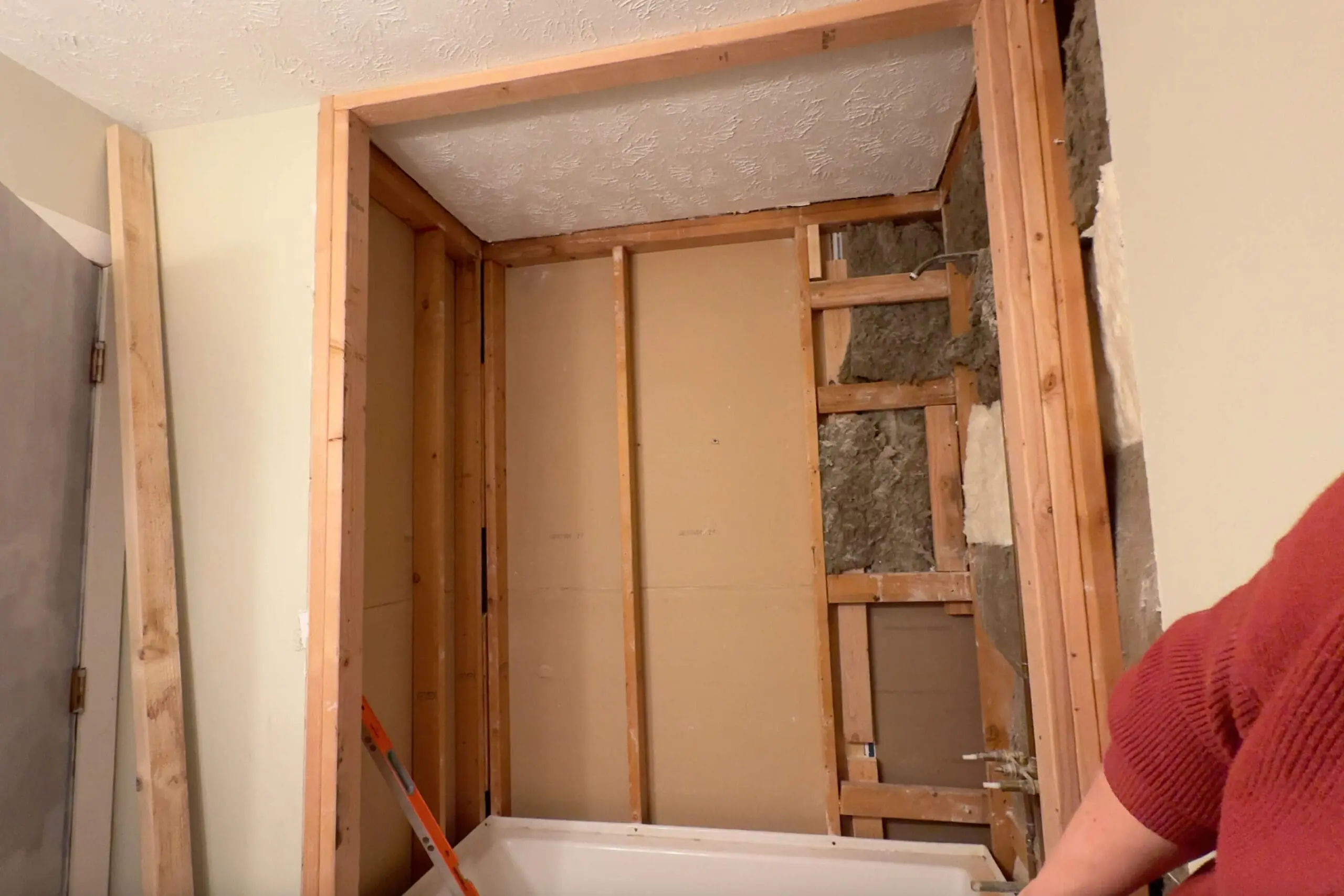 An in-progress bathroom remodel showing a fully framed wall around a white bathtub. The framing reveals studs, insulation, and partially exposed drywall, with a ladder and tools nearby.